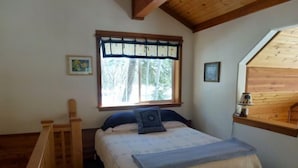 A bedroom with a bed, a wooden headboard, a window with a view of trees, and a wooden desk with a lamp.