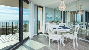 A balcony with a view of the beach, a dining area with white chairs and a round table, and two chandeliers.