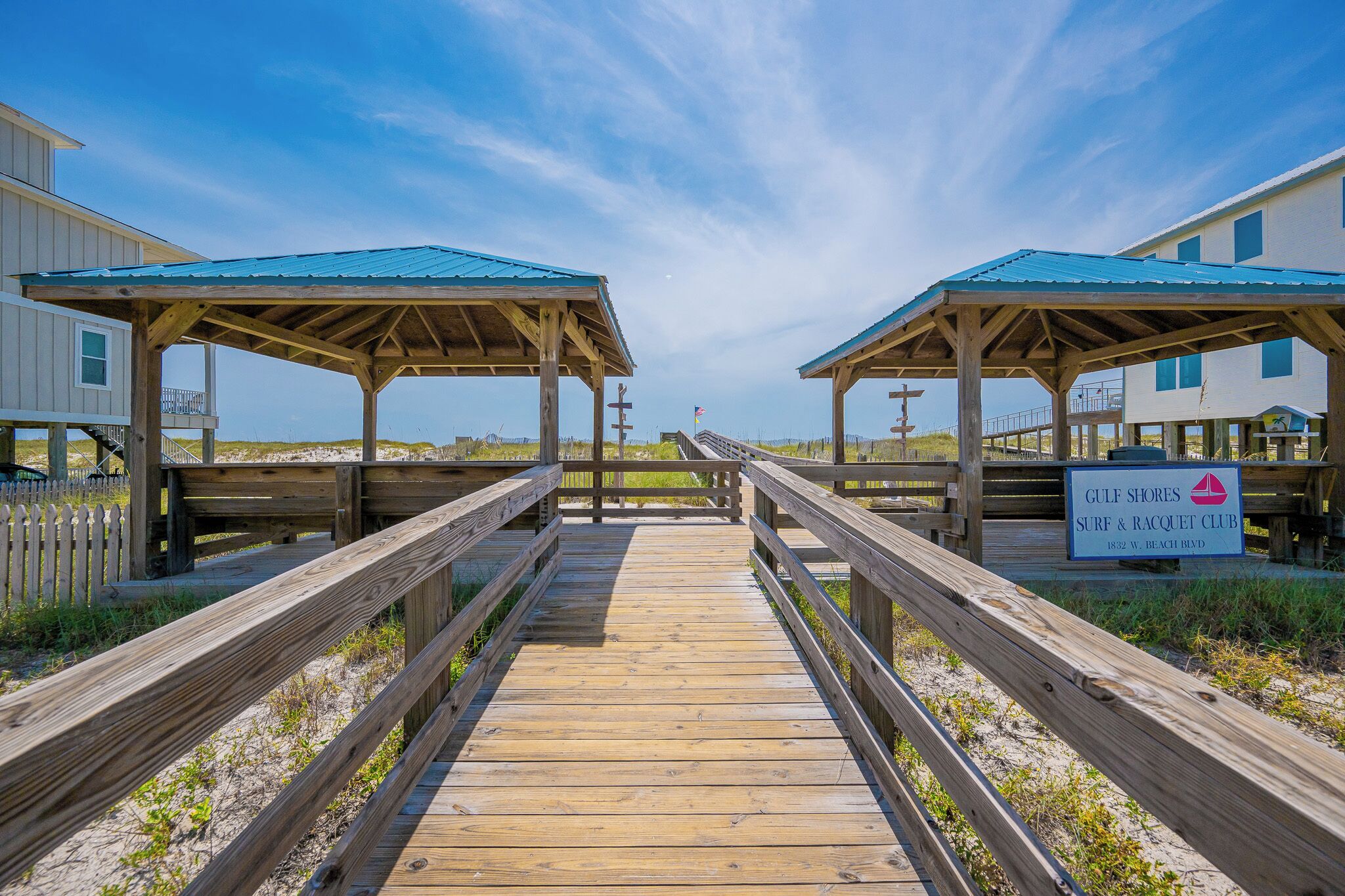A wooden boardwalk leading to beach huts with a sign for Gulf Shores Surf & Racquet Club.