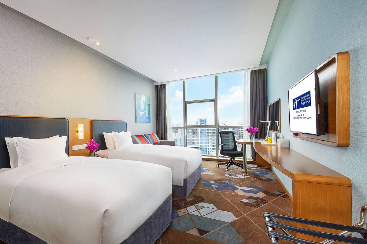 Ein Hotelzimmer mit zwei Betten, einem Schreibtisch, einem Stuhl, einem großen Fenster mit Blick auf die Stadt und einem Bildschirm, der Hotelinformationen zeigt.