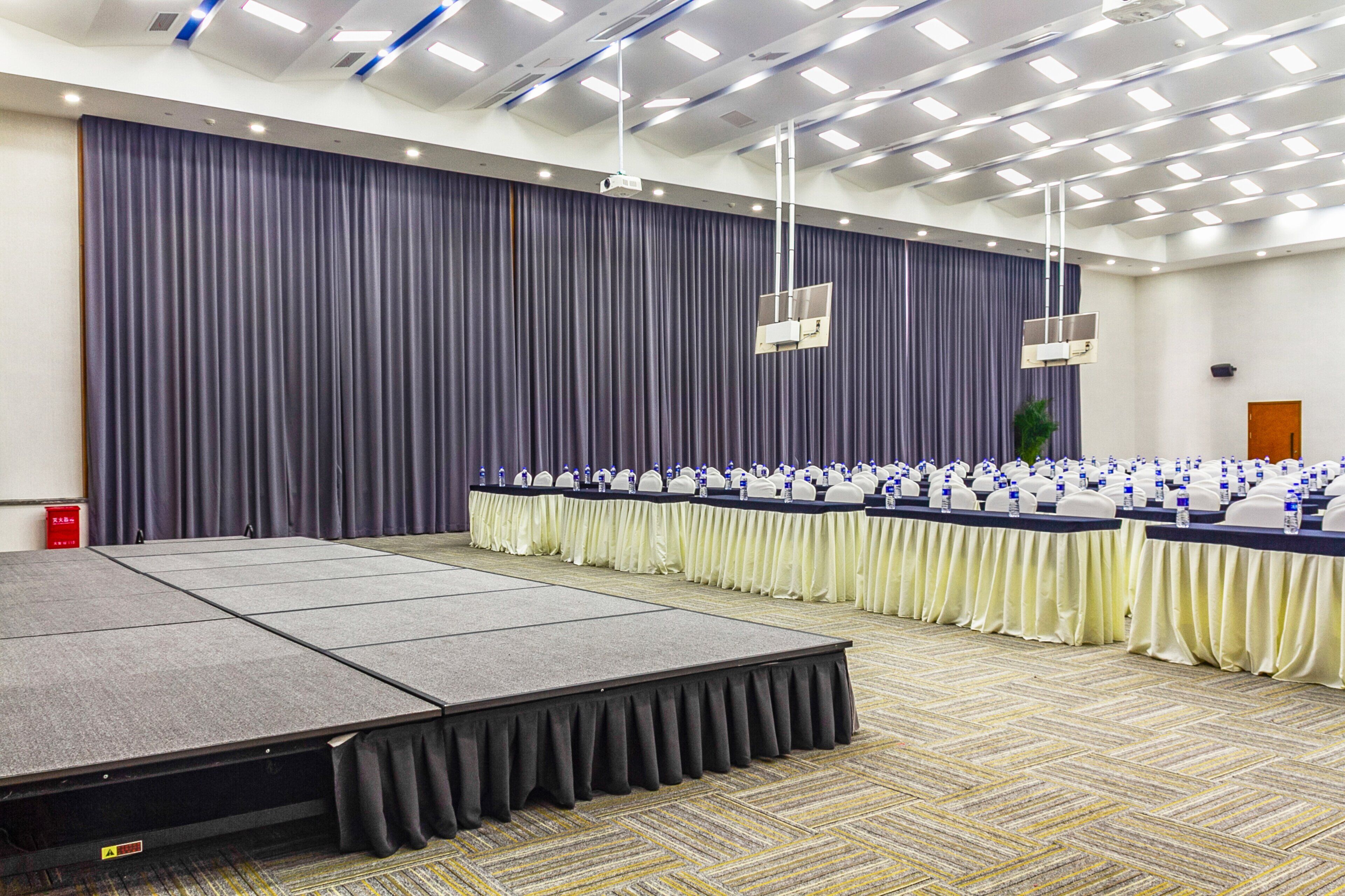 Une salle de conférence avec une scène, des rangées de tables et de chaises, et un grand rideau en fond.