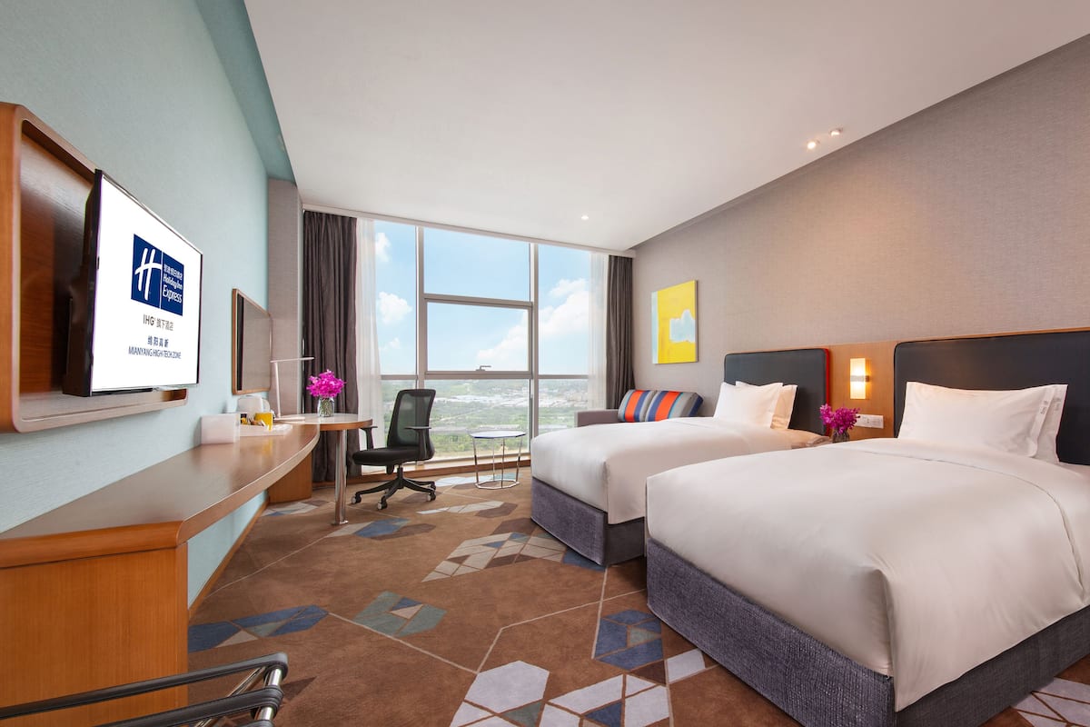 Ein Hotelzimmer mit zwei Betten, einem Schreibtisch, einem Sessel und einem großen Fenster mit Blick auf die Stadt.