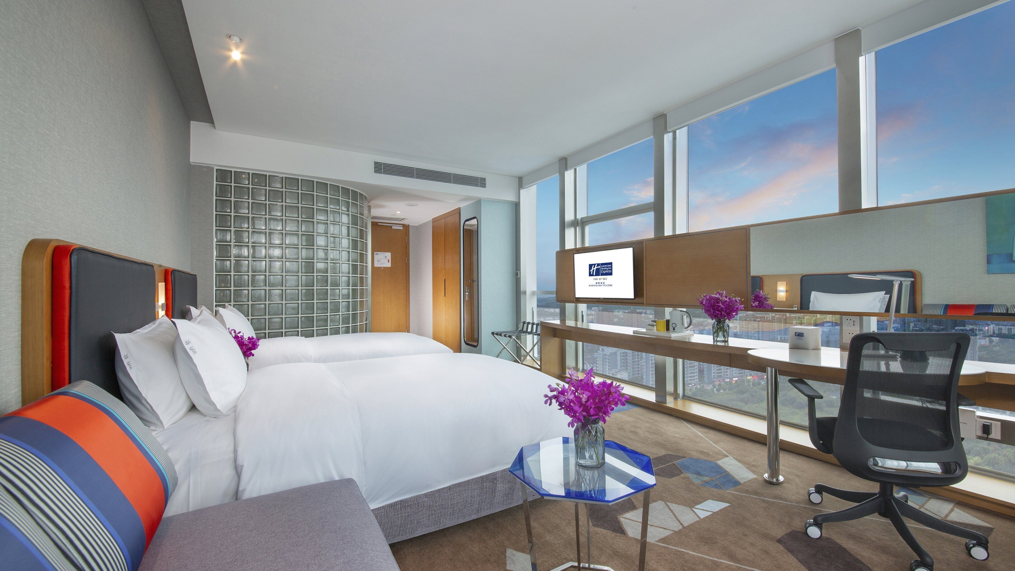 Une chambre d’hôtel avec deux lits, un bureau, une chaise, une petite table et une grande fenêtre donnant sur la ville.