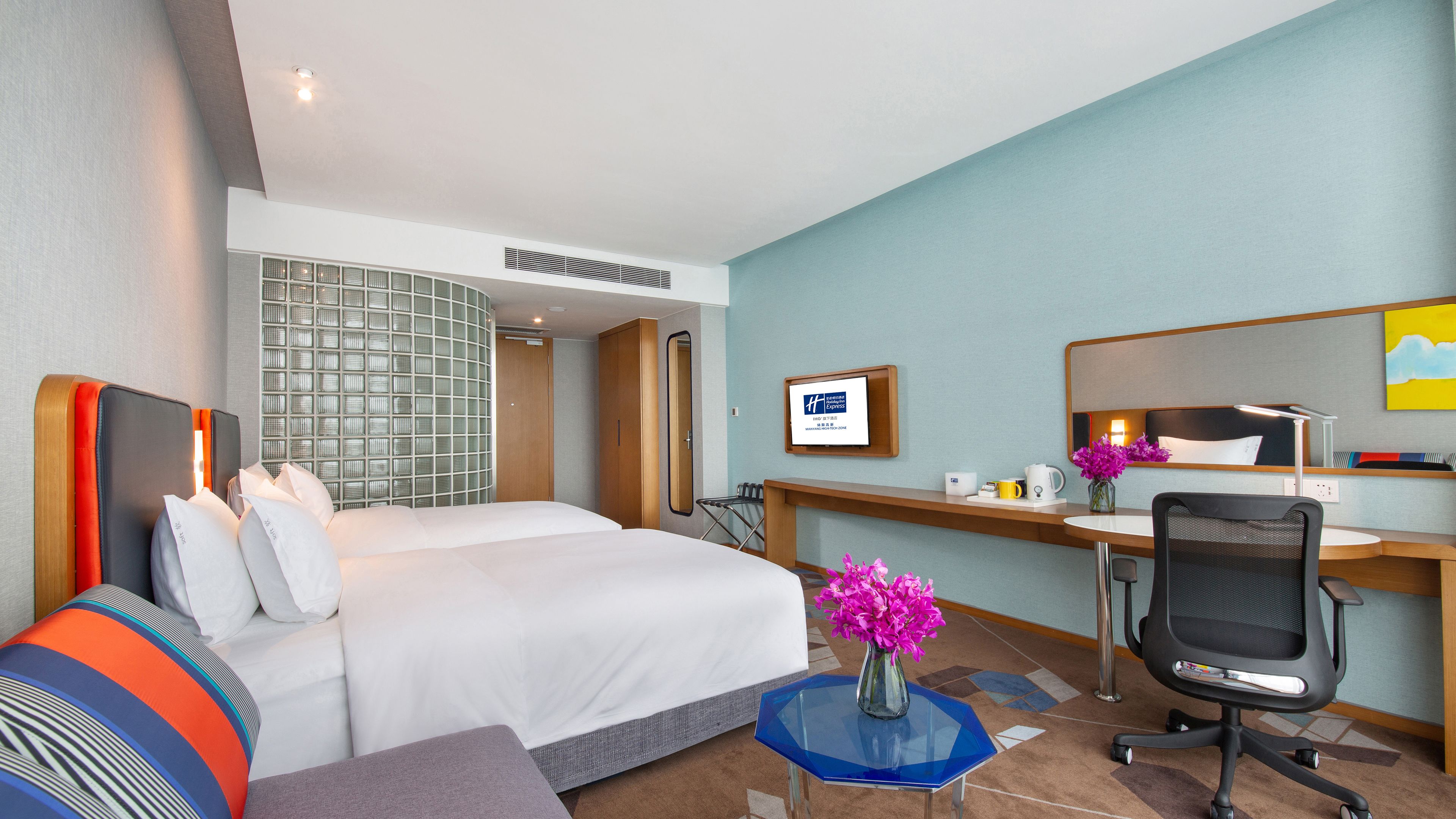 A hotel room with a bed, a desk, a chair, a small table, and a mirror.