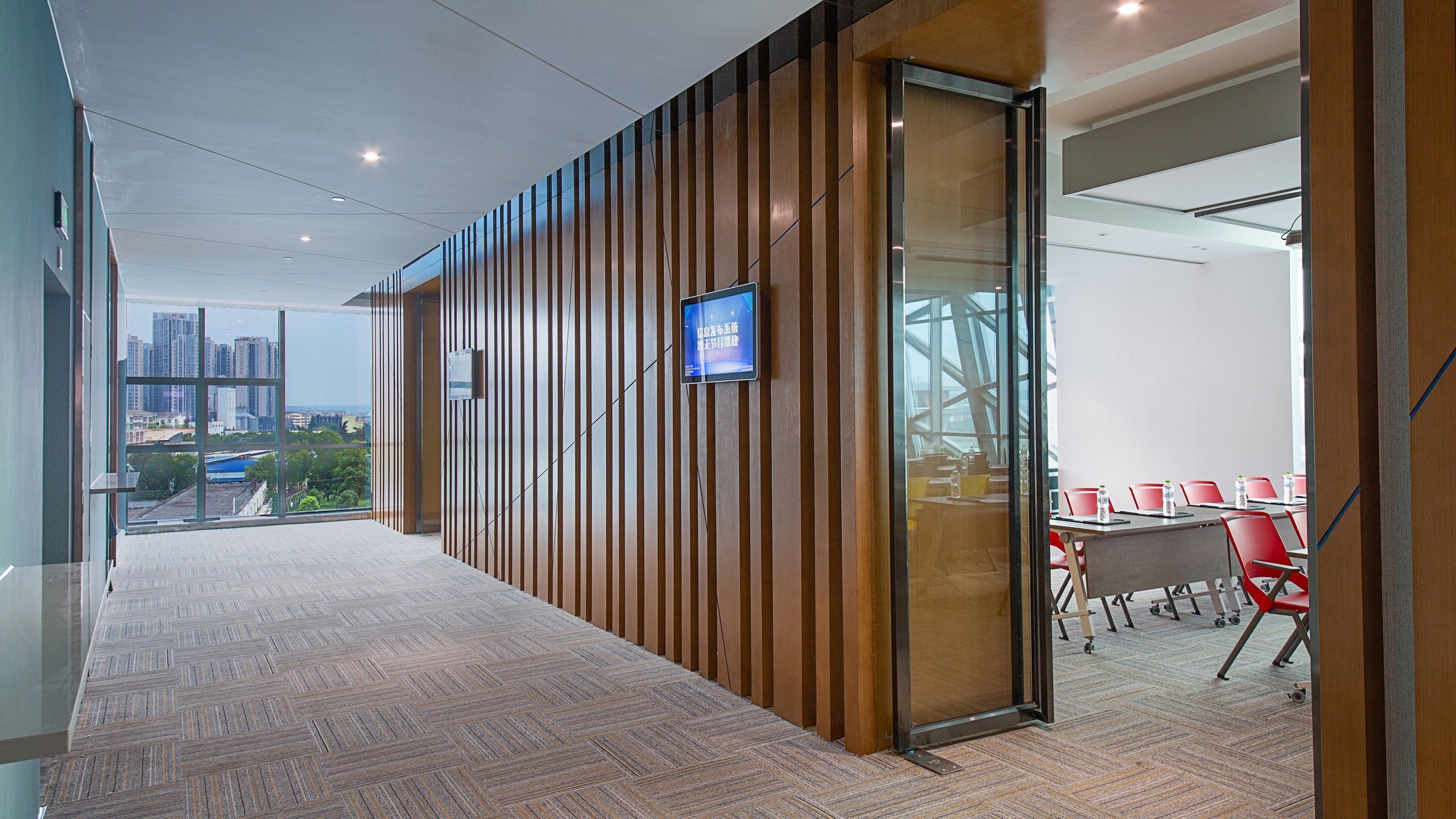 A modern conference room with a large glass wall, a digital screen, and a view of the cityscape.