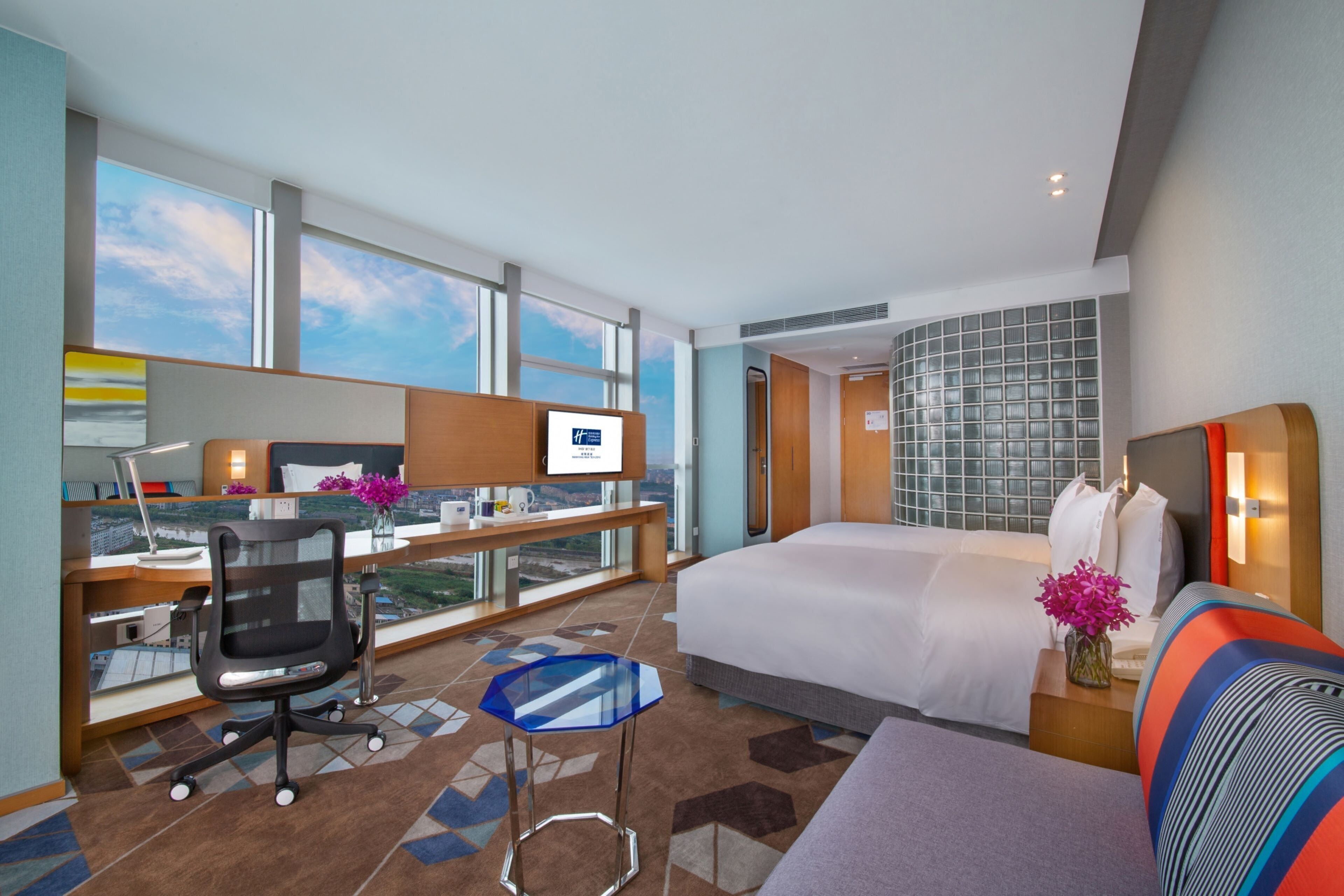 A hotel room with a large bed, a desk with a computer, a chair, a small table, and a view of the city.