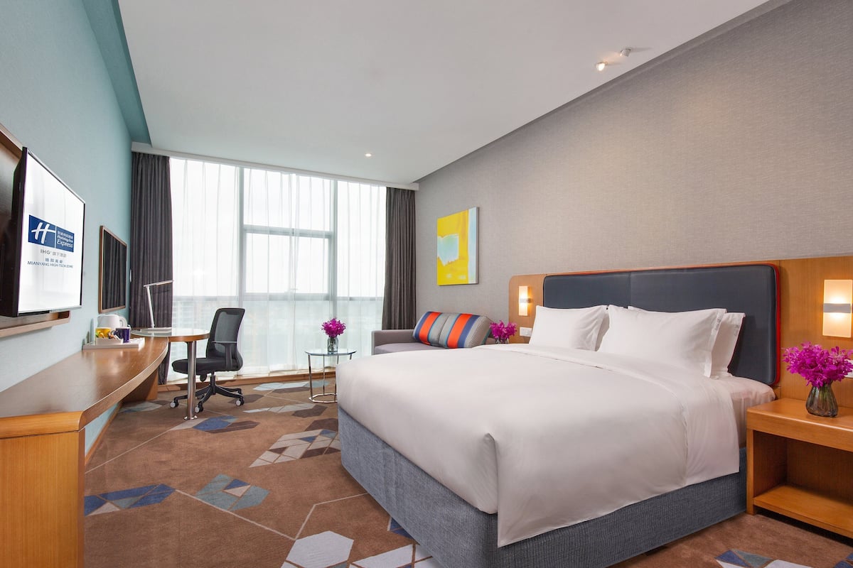 Ein Hotelzimmer mit einem großen Bett, einem Schreibtisch mit Computer, einem Sessel und einem Fernseher an der Wand.