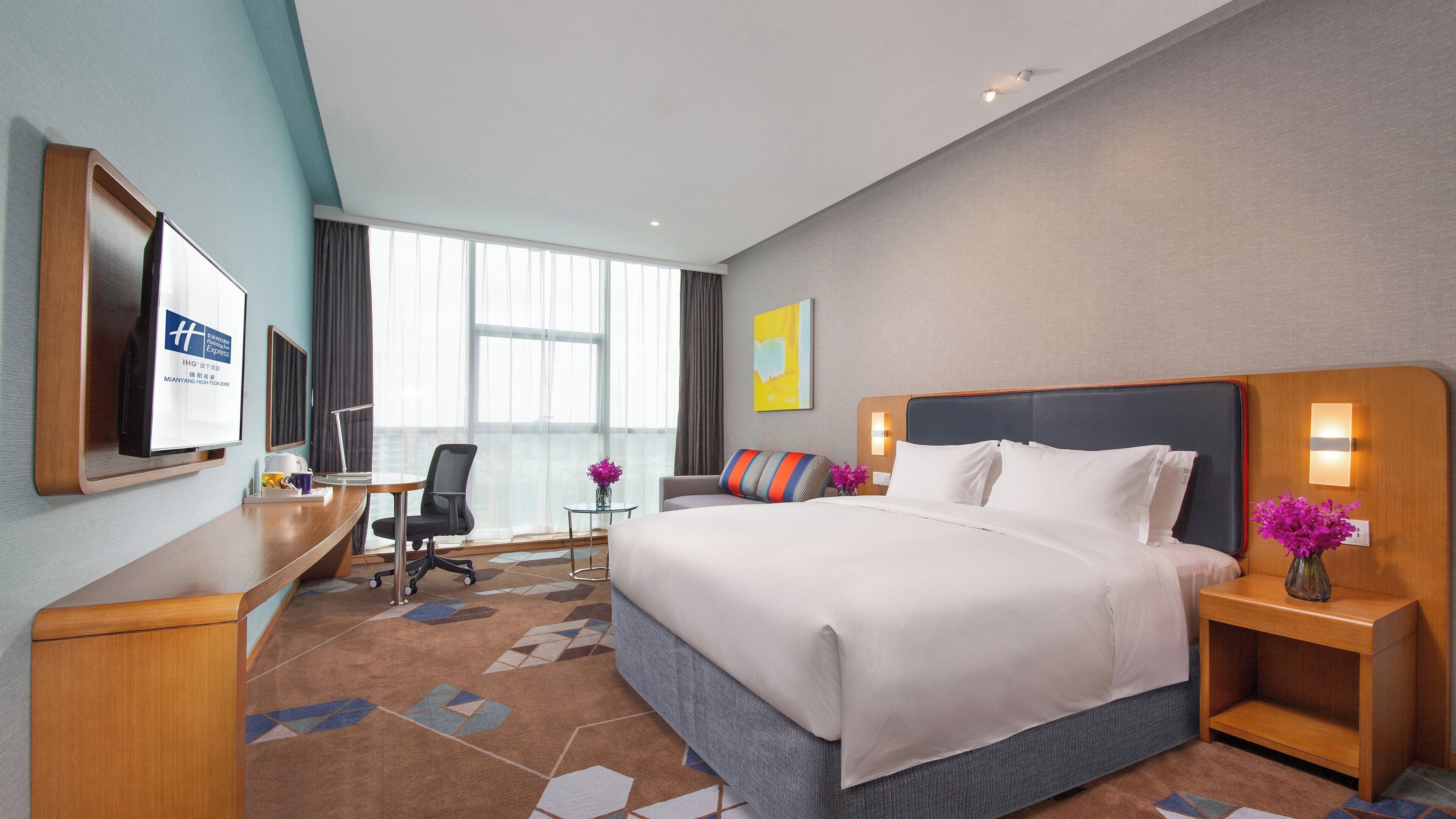 Ein Hotelzimmer mit einem großen Bett, einem Schreibtisch, einem Sessel, einem Fernseher und einem Fenster mit Vorhängen.