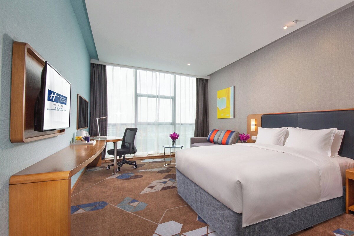 A hotel room with a large bed, a desk with a computer, a chair, and a TV on the wall.