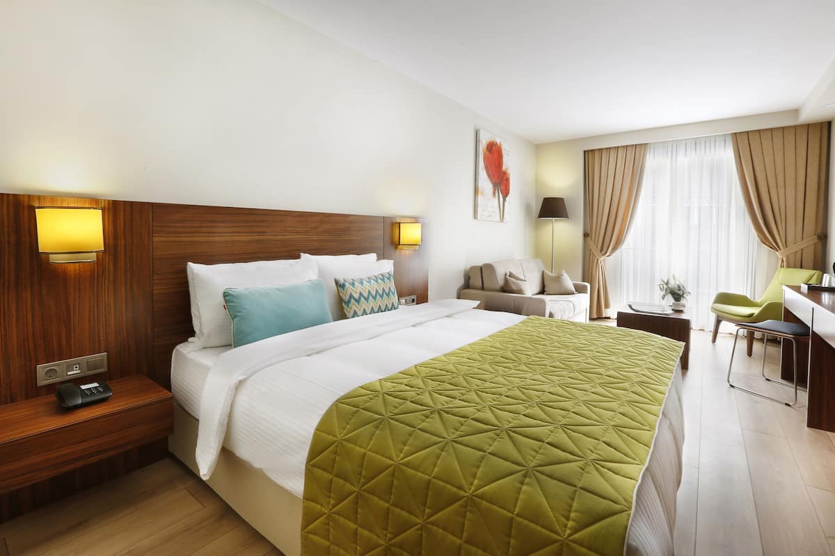 A hotel room with a wooden headboard, a green quilted bedspread, a desk with a chair, and a window with curtains.