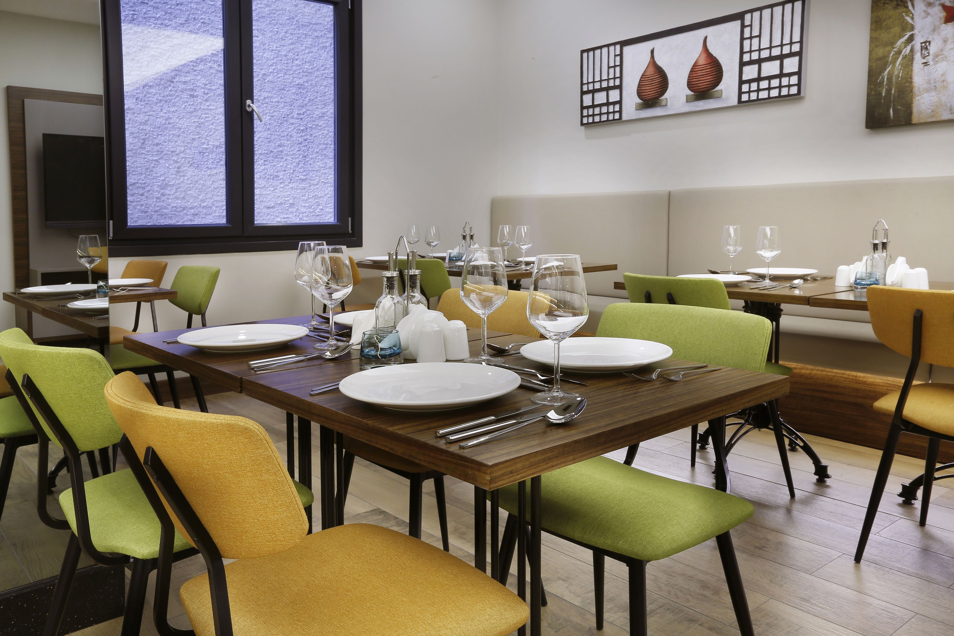 A dining area with multiple tables set for two, each with green and yellow chairs, white plates, and wine glasses.