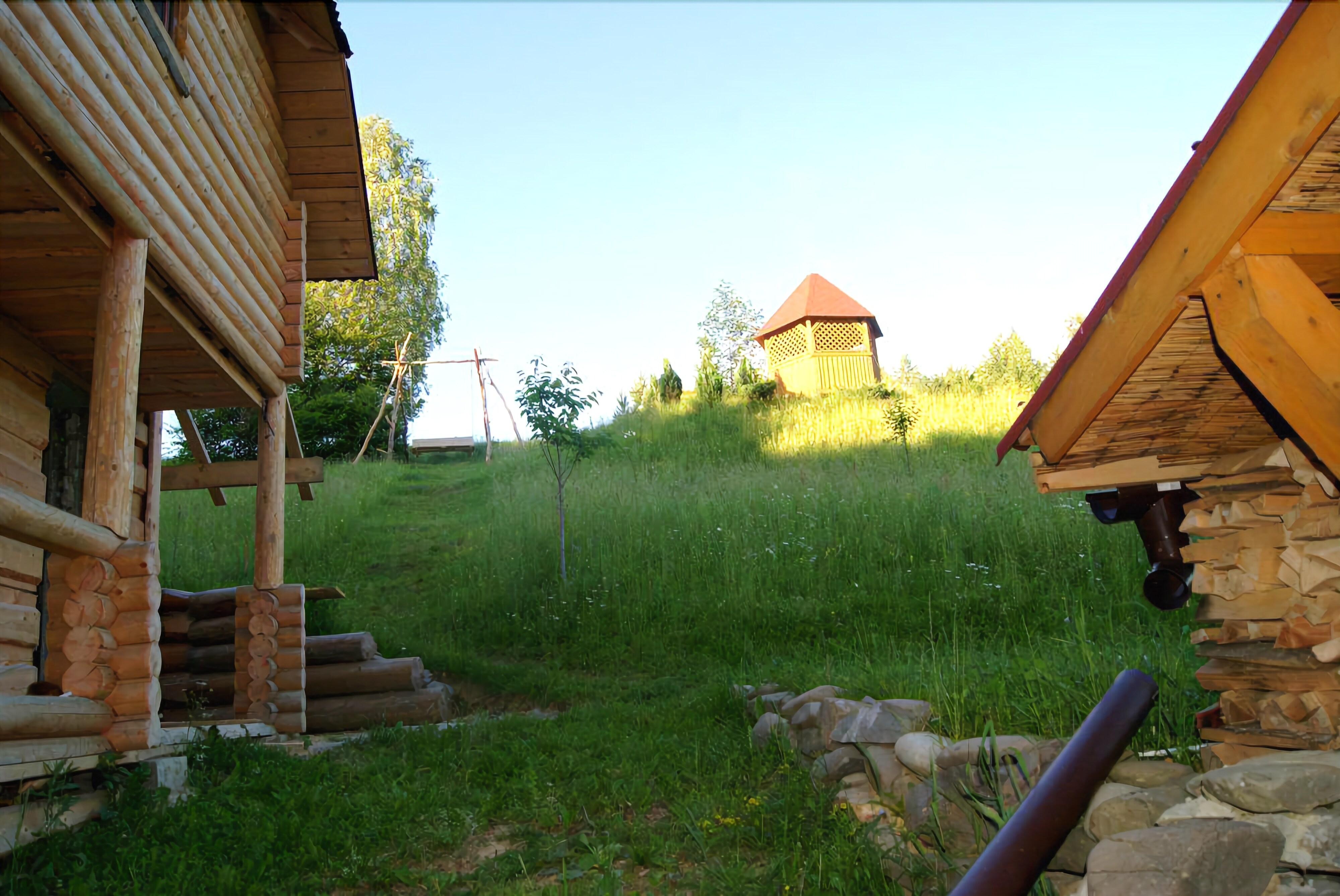 A rustic wooden cabin with a stone foundation, a small wooden structure in the background, and a grassy area with a stone wall.