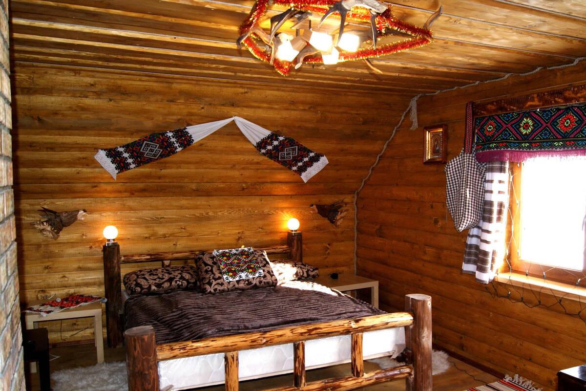 A cozy log cabin room with a wooden bed, two bedside lamps, a patterned blanket, and a decorative wall hanging.