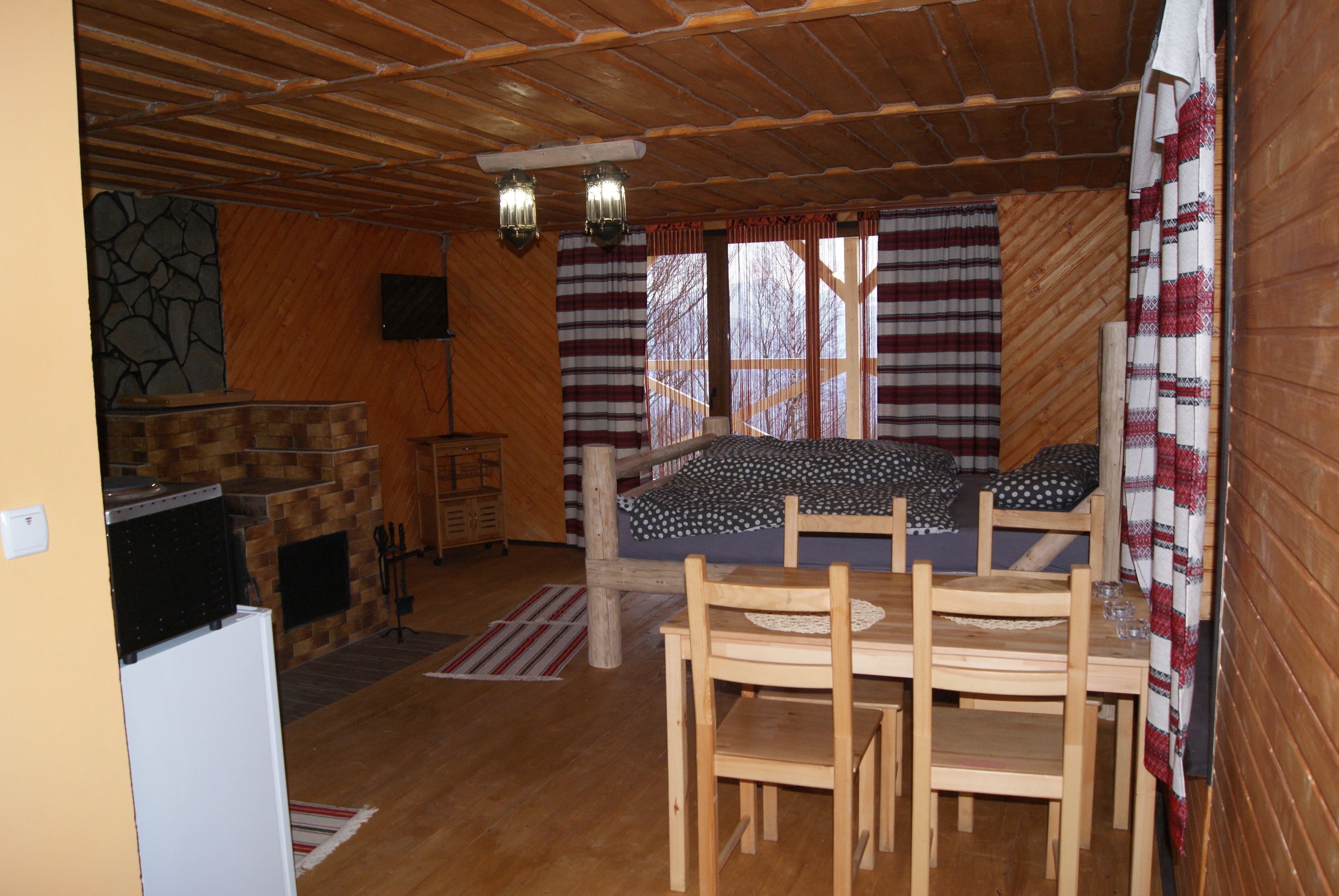 A cozy cabin interior with a wooden ceiling, a bed with patterned bedding, a dining table with chairs, and a small kitchen area with a refrigerator.