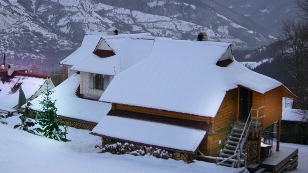 Een huis bedekt met sneeuw met een houten trap naar de ingang, omgeven door bergen en een zonsonderganglucht.