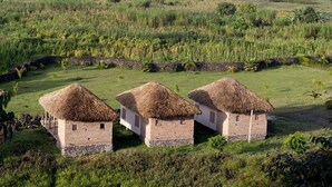 Three thatched-roof huts with stone bases, surrounded by lush green fields and a body of water in the background. - Lake Chahafi  Resort (Kisoro)