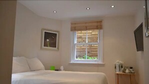 Un dormitorio con cama, una ventana con persianas, una mesita de noche con hervidor y un cuadro en la pared.