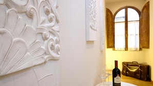 A room with a white wall featuring a decorative plaster design, a wooden window with curtains, and a table with a bottle of wine and two glasses.