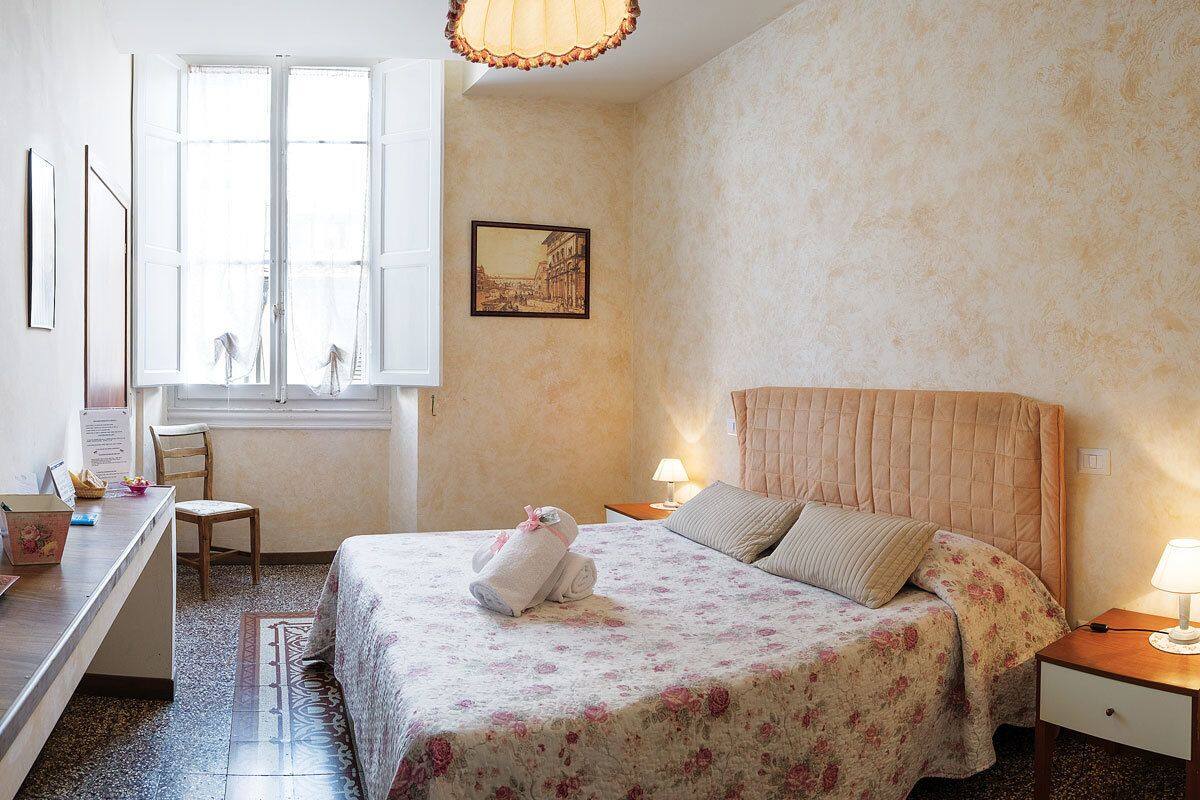 Een slaapkamer met een bed, een bureau, een stoel, een raam met gordijnen, een bedlampje en een lijst aan de muur.
