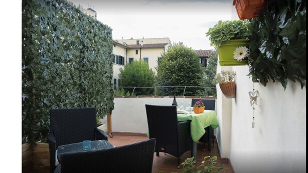 A narrow outdoor corridor with a high wall on one side and a white wall on the other. There are two outdoor seating areas with tables and chairs, and a potted plant is visible on the right side.
