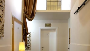 A narrow hallway with a round table and two chairs, a bench, a lamp, a door, and a window with a curtain.