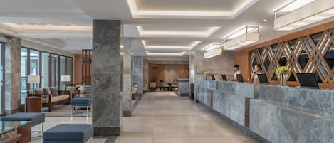 A modern hotel lobby with a reception desk, seating area, and a view of the city.