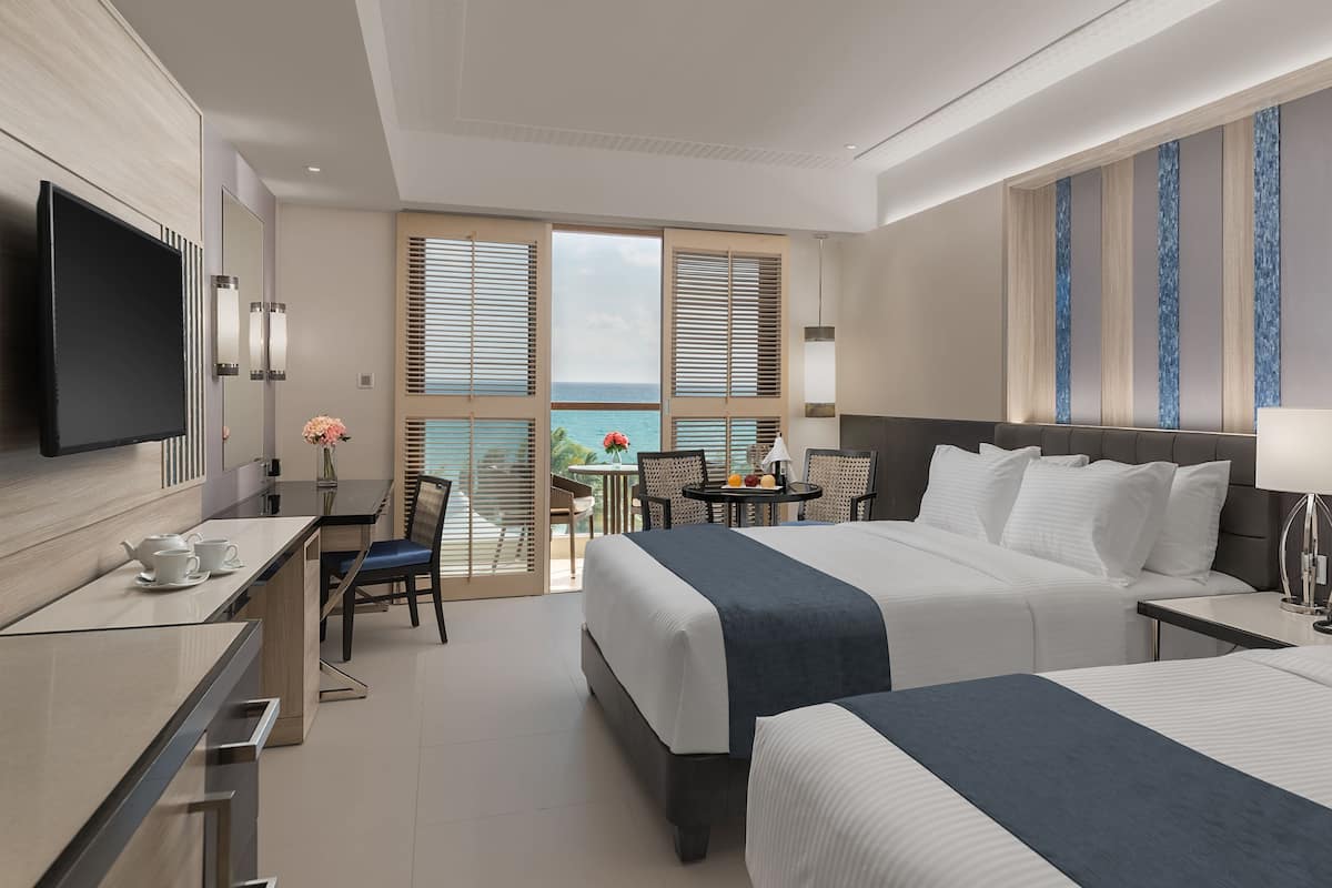 Habitación de hotel con dos camas, un escritorio con televisor, una mesita con sillas y vista al océano.