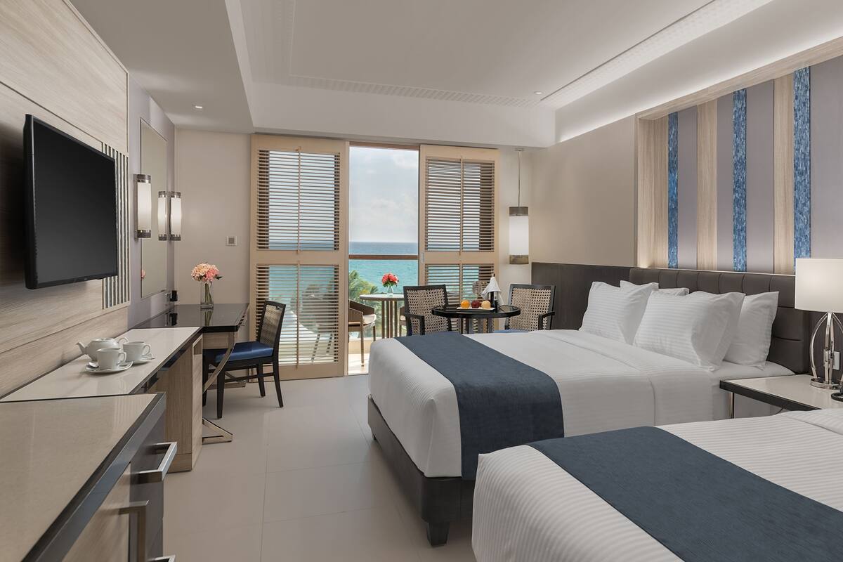Habitación de hotel con dos camas, un escritorio con televisor, una mesita con sillas y vista al océano.