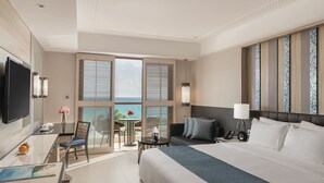 Een hotelkamer met een bed, een bureau met stoel, een televisie en een balkon met uitzicht op zee.