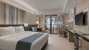 Een moderne hotelkamer met een groot bed, een bureau, een televisie en een balkon met uitzicht op palmbomen.