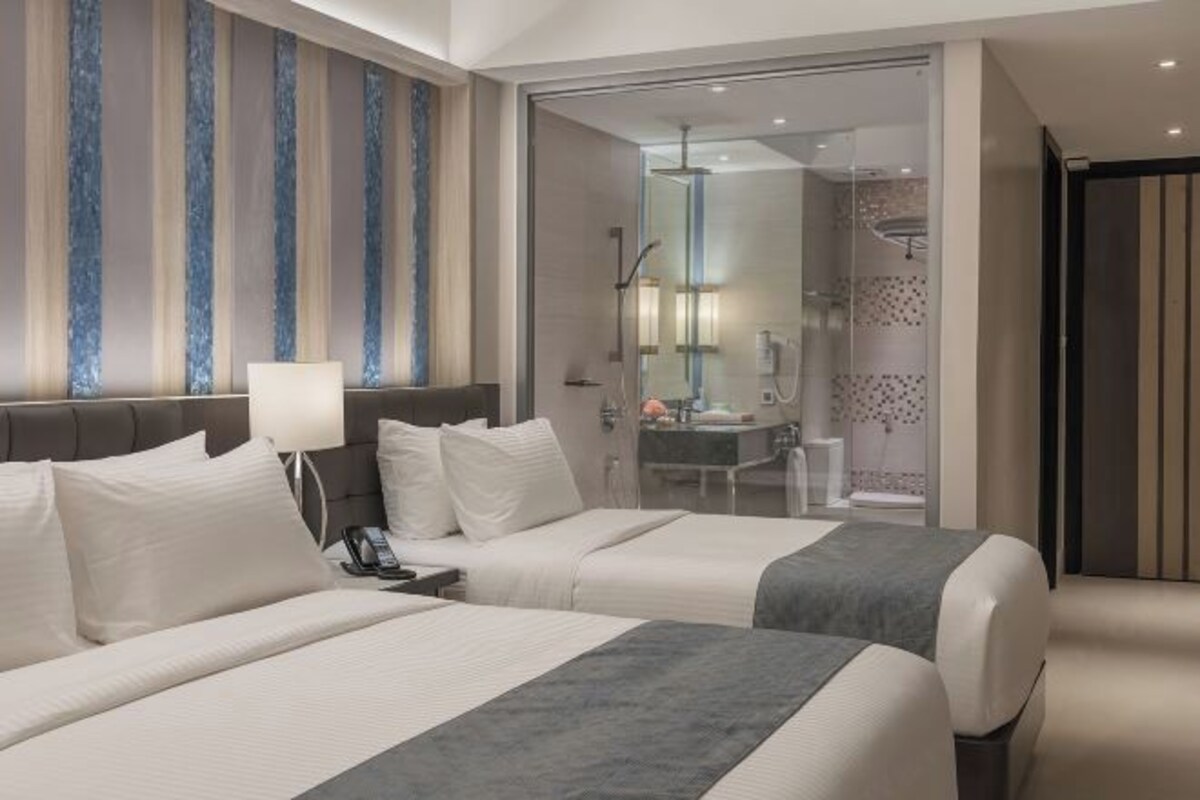 Une chambre d’hôtel avec deux lits, une table de chevet avec une lampe, une salle de bain avec une douche et des toilettes visibles à travers une porte en verre.