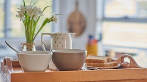 Ein Holztablett mit einer weißen Schüssel, einer braunen Schüssel, einer weißen Kanne mit einem Löffel, einem Glas mit grünen Kräutern und Brotscheiben.