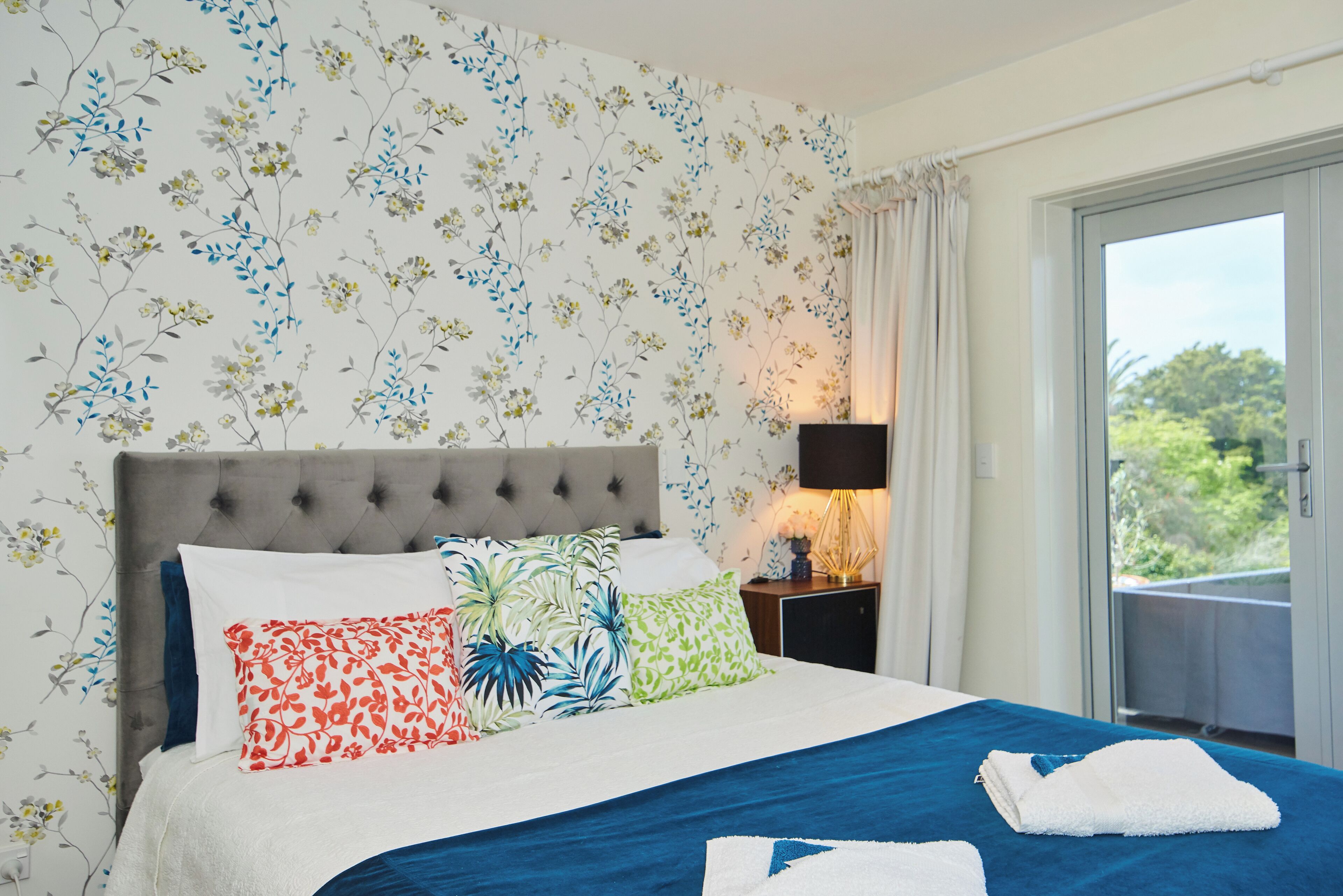 A hotel room with a bed, floral wallpaper, a nightstand with a lamp, and a view of greenery through a sliding door.