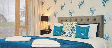 A bedroom with a large bed, blue and white bedding, a nightstand with a lamp, and floral wallpaper.