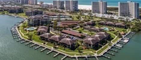 An aerial view of a coastal resort with multiple buildings, a marina, and a beach in the background.
