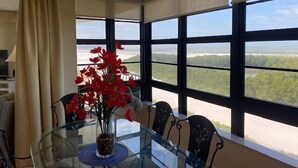 A dining area with a glass table, red flowers, and a view of a forest through large windows. - 2/2 condo, with fantastic views of the beach (Marco Island)