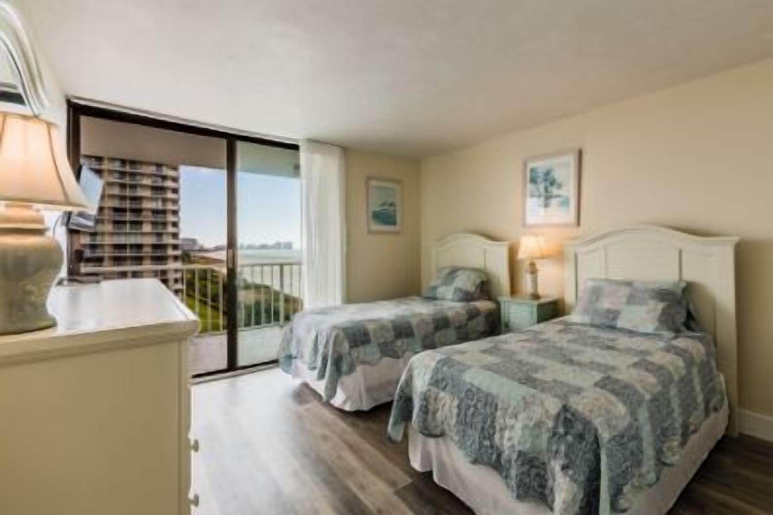 Un dormitorio con dos camas, una mesita de noche, una lámpara y vistas a un balcón con un edificio al fondo.