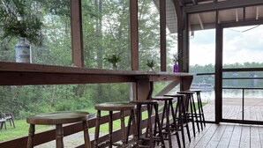 Un espacio de estar al aire libre con techo, con bancos y mesas de madera, con vista a una masa de agua y rodeado de árboles.