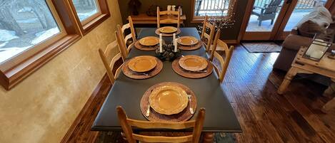 A dining area with a table set for six, wooden chairs, and a view of the outdoors through large windows.