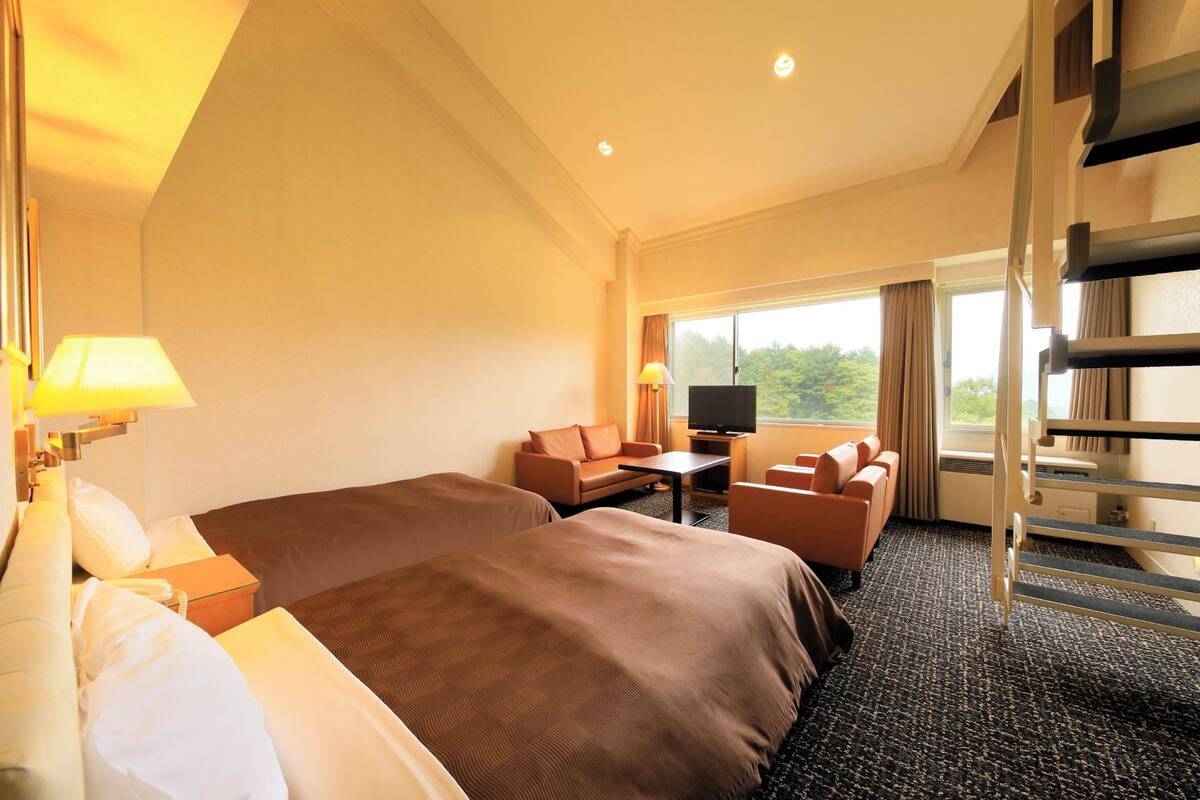 A hotel room with two beds, a sofa, a desk, a TV, and a window with a view of greenery.
