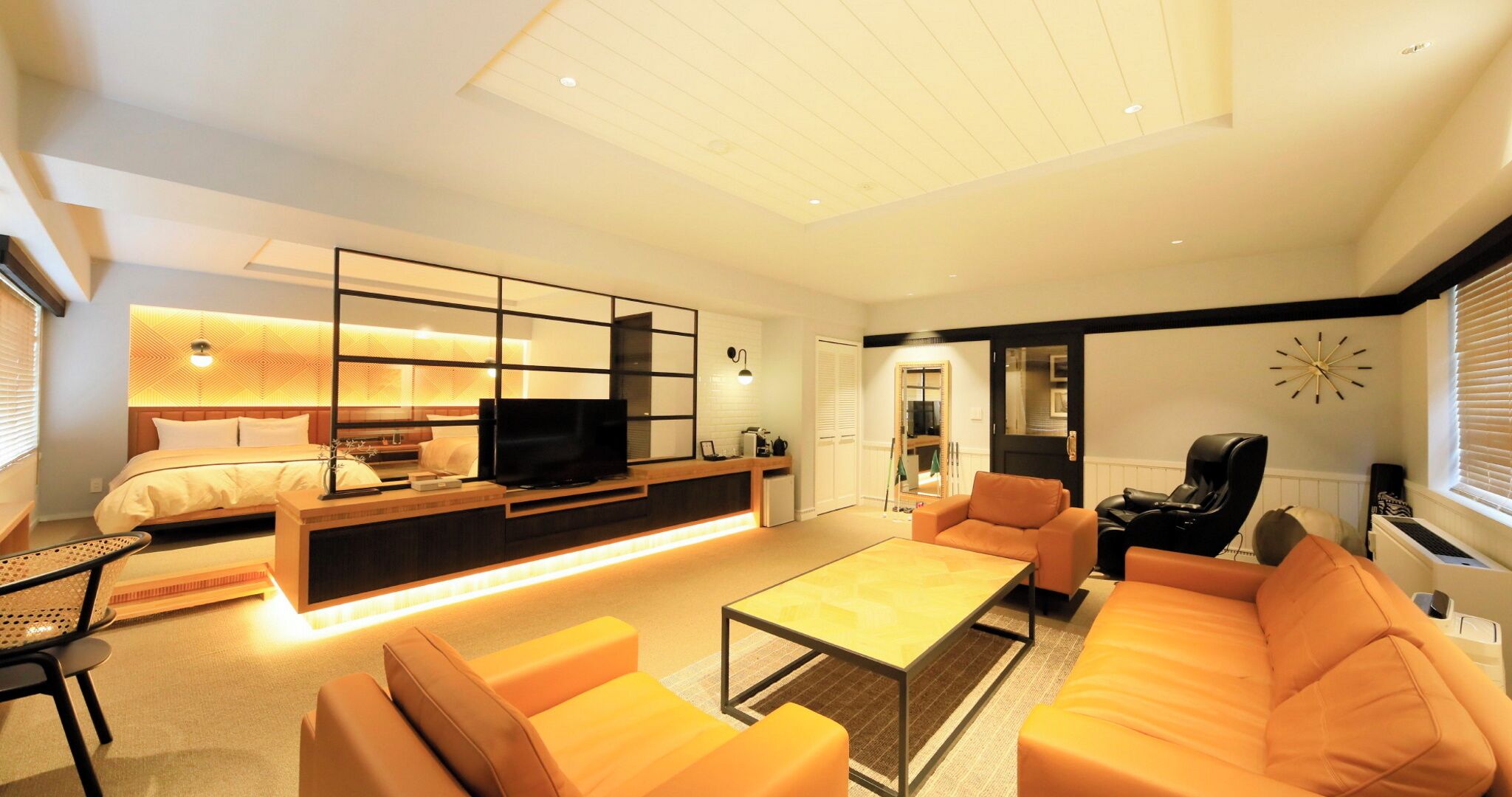 A modern living room with a sofa, coffee table, and TV. There is a bed with a headboard and a small table beside it.