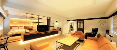 A modern living room with a sofa, coffee table, and TV. There is a bed with a headboard and a small table beside it.