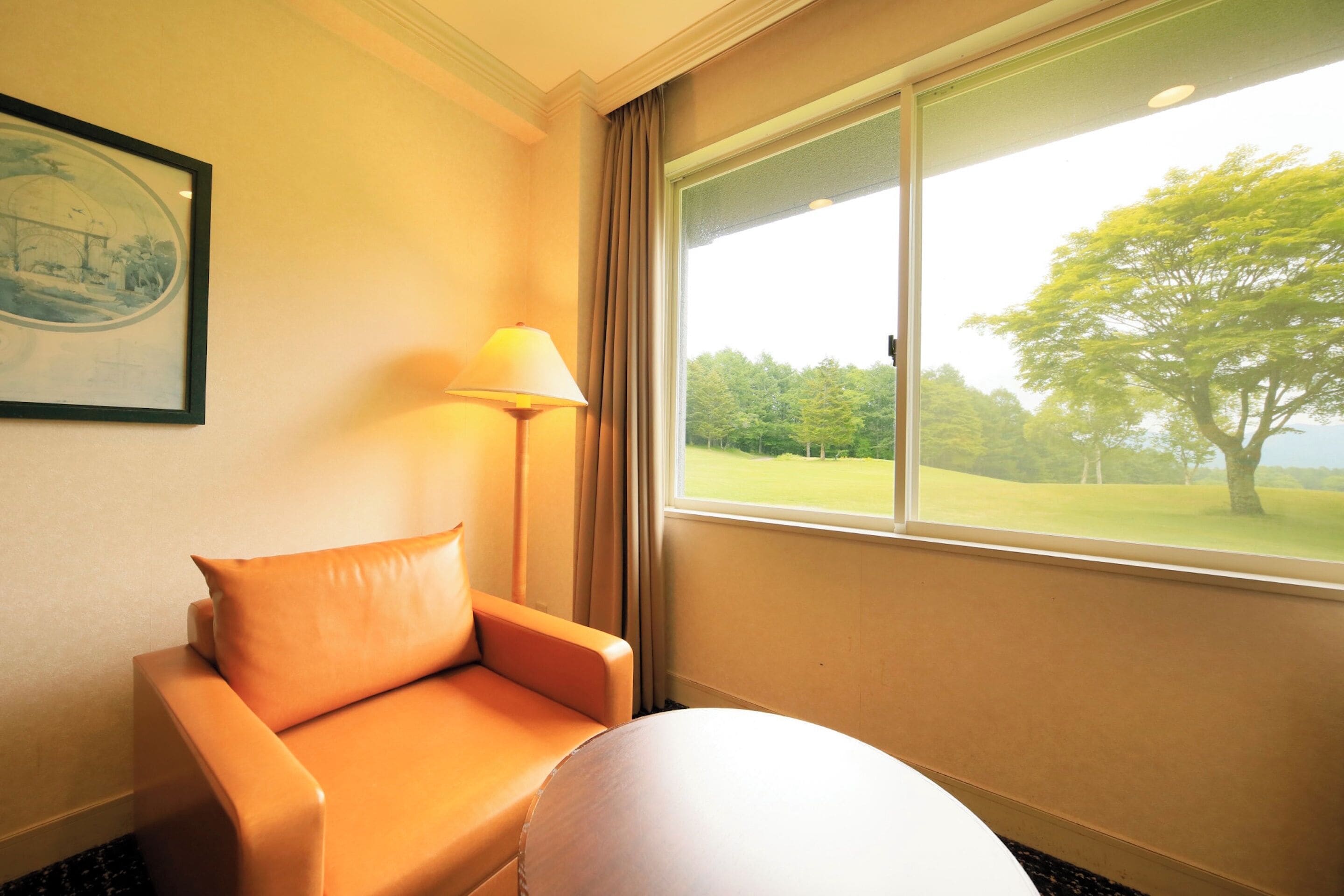A room with a beige sofa, a round table, a framed picture on the wall, and a window with a view of a tree and a golf course.