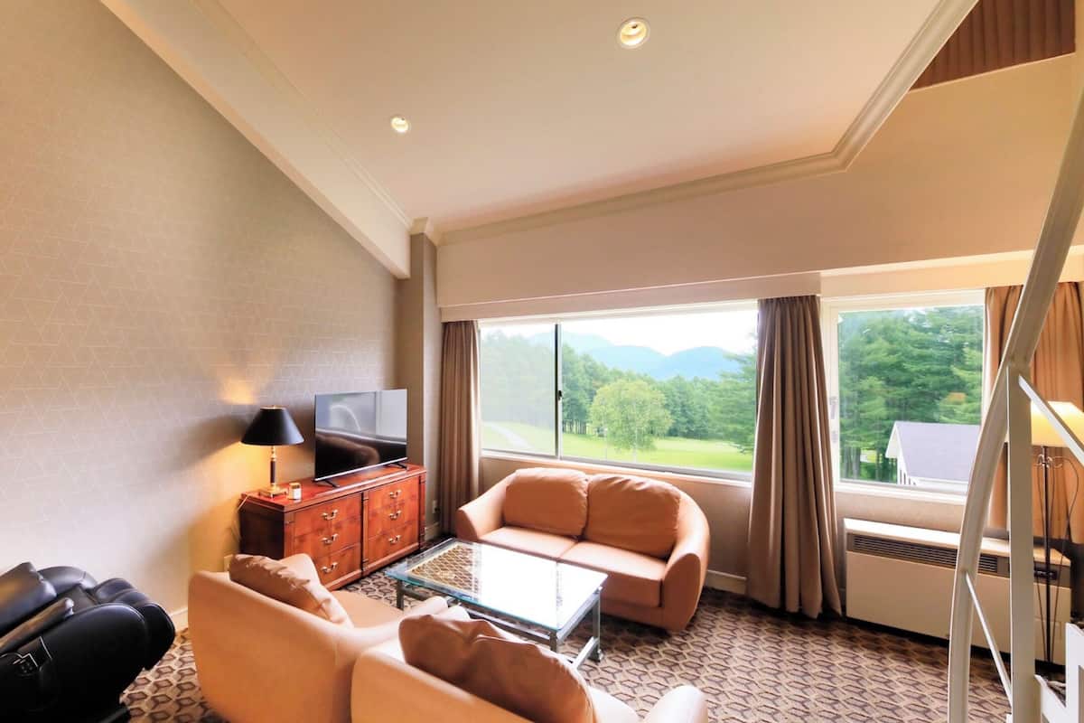 A hotel room with a sofa, armchair, and a glass coffee table. There is a TV on a wooden cabinet, and a window with a view of greenery.