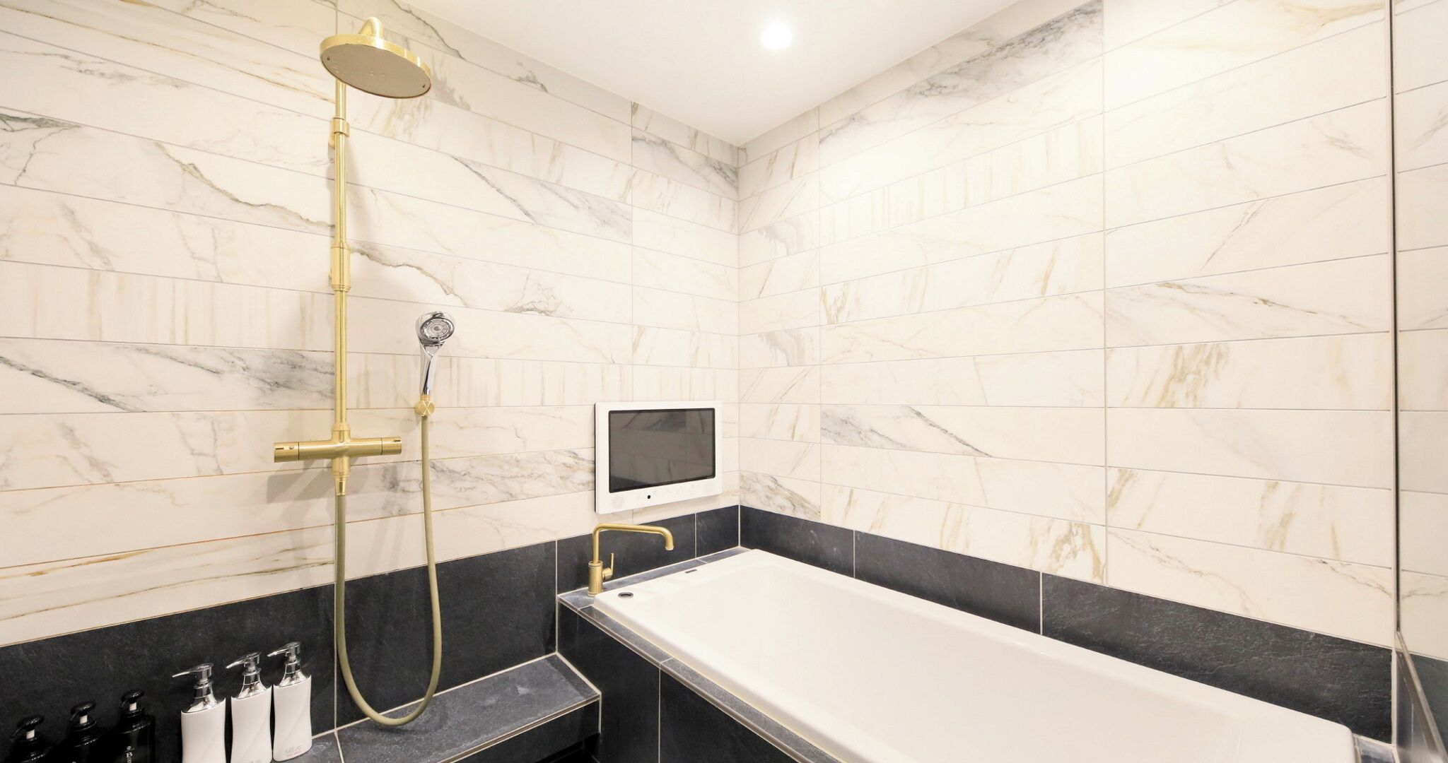 A bathroom with a bathtub, a handheld showerhead, a wall-mounted TV, and a row of toiletries.