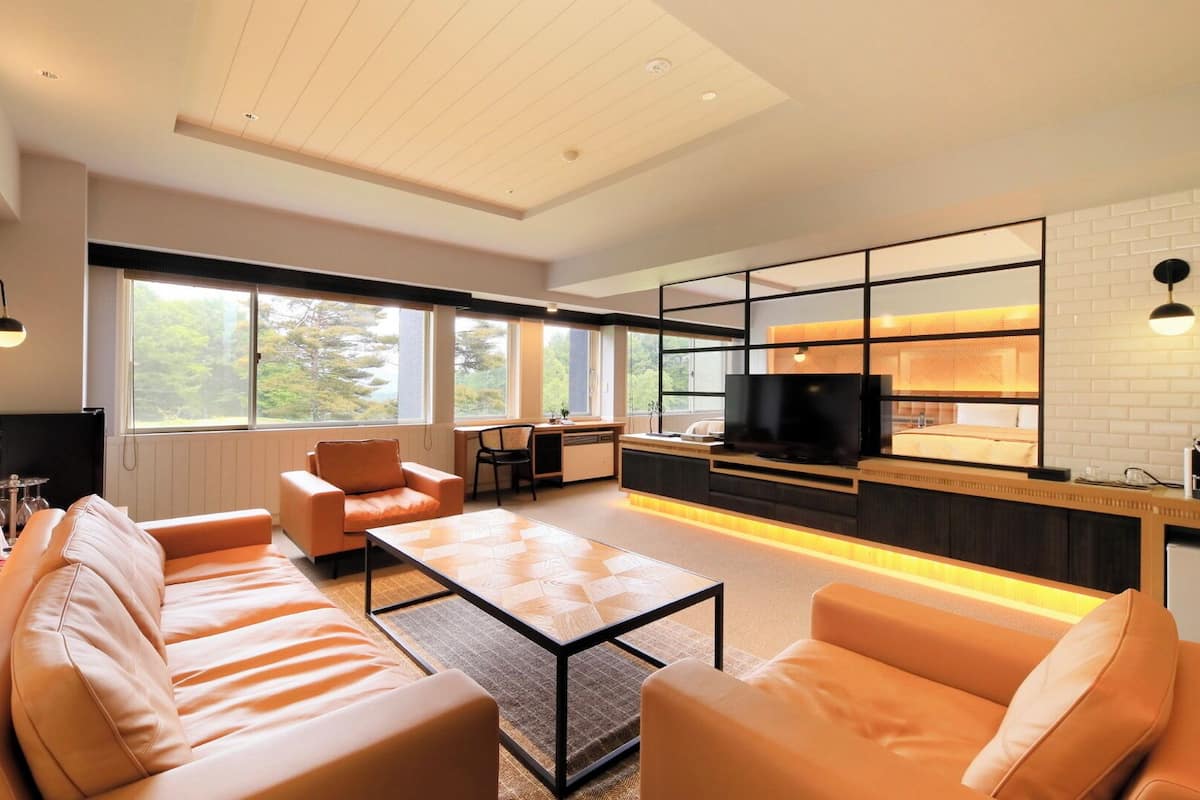 A modern living room with a sofa, coffee table, and TV. There is a kitchen area with a counter and appliances. Large windows provide natural light and views of the outdoors.