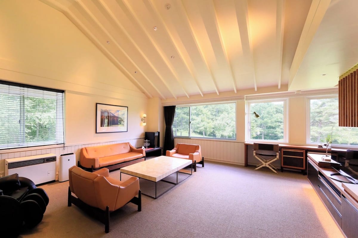 A modern living room with a sofa, coffee table, and exercise equipment. Large windows provide natural light and views of greenery.