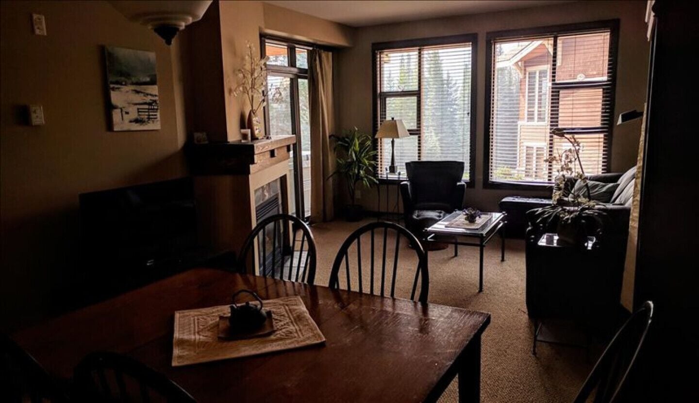 Un salon chaleureux avec une table à manger en bois, des chaises et un canapé. De grandes fenêtres laissent entrer la lumière naturelle et offrent une vue sur l’extérieur.