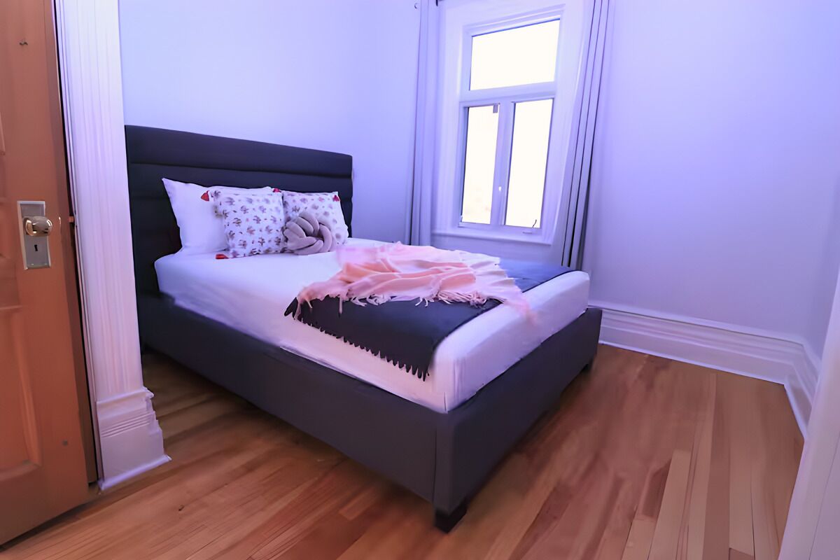 A neatly made bed with a dark headboard, white bedding, and a pink blanket. A window with white curtains is visible to the right of the bed.
