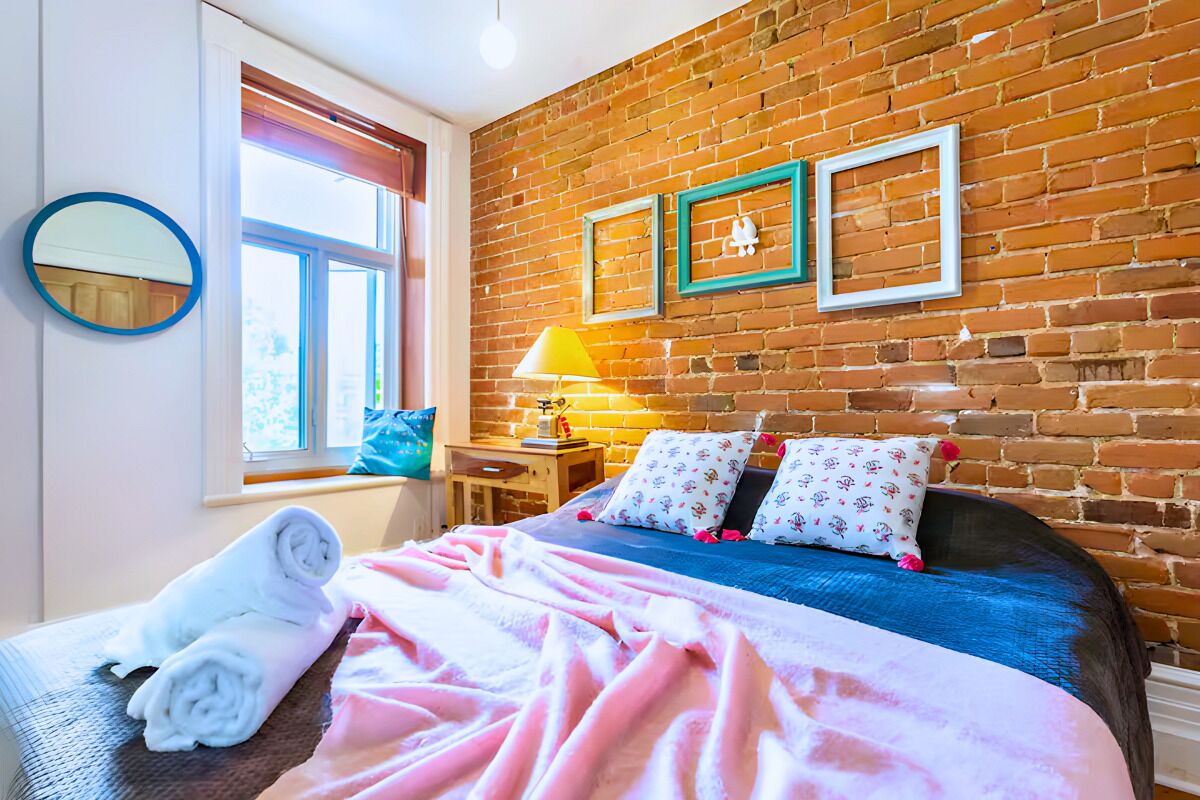 A bedroom with a brick wall, a bed with a blue bedspread, two pillows, a bedside table with a lamp, a window with curtains, and a round mirror on the wall.